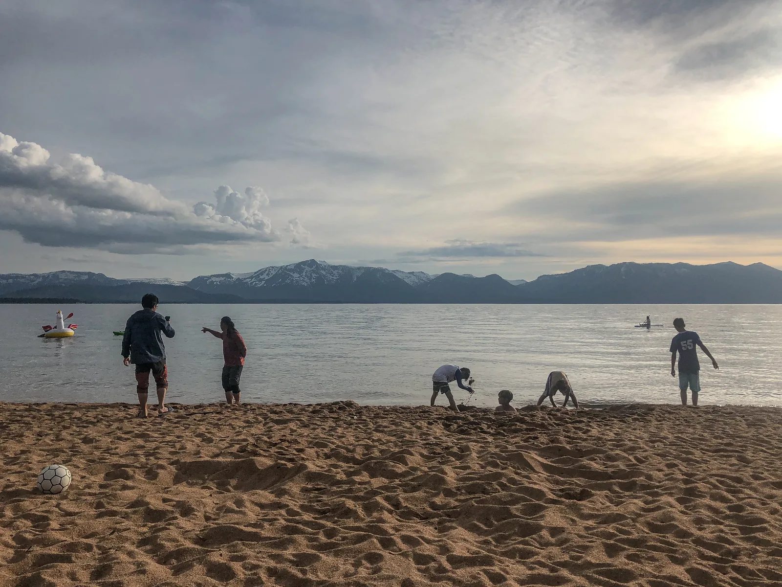 湖畔沙地上有人站立、蹲下或坐著，遠處是山巒。