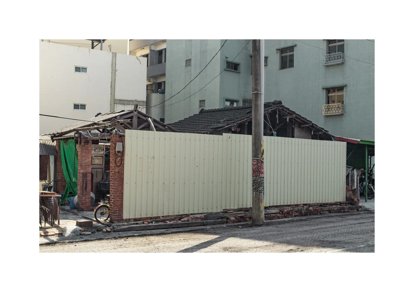 小冊子《B》第 27 頁：白色鐵皮圍籬遮住低矮磚屋，部分屋頂骨架露出，後方是住宅大樓。
