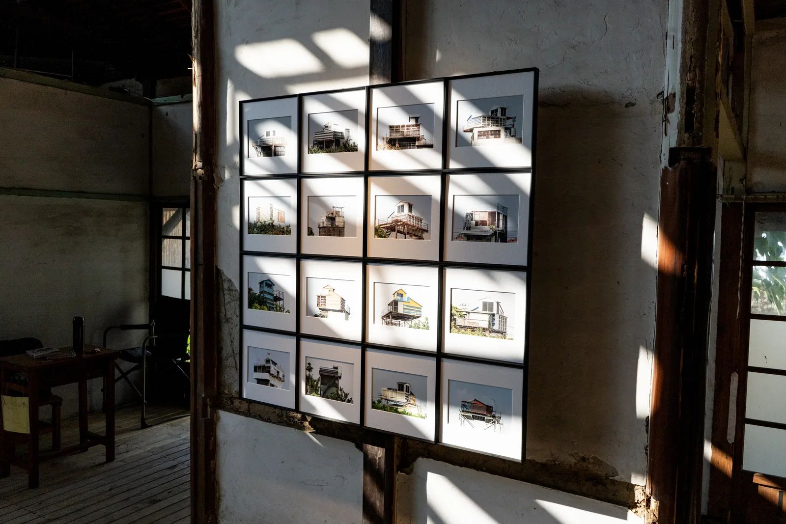 An angled view of the four-by-four pigeon-loft display shows window shadows across its white mounts, with wooden flooring and furniture nearby.